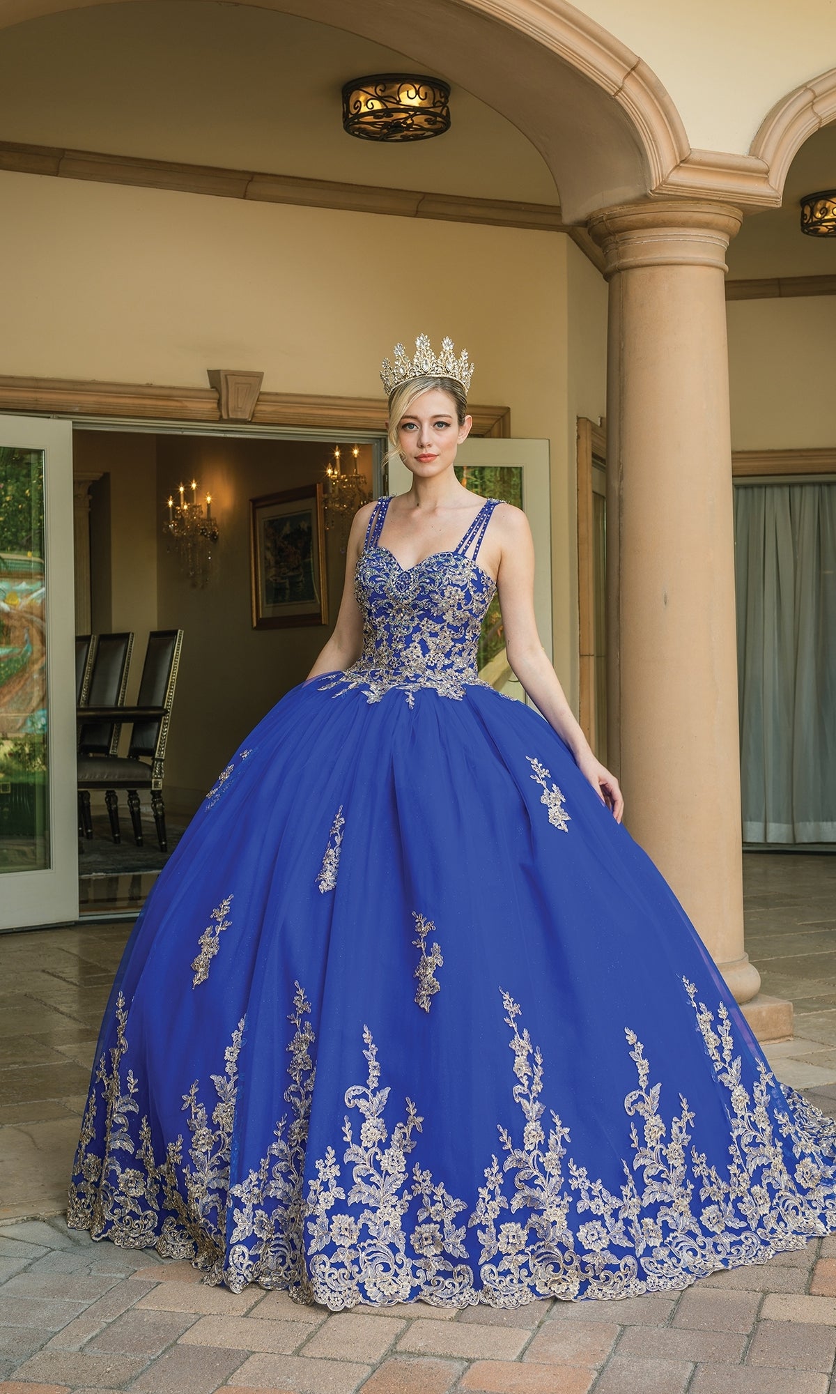 Wearing the Royal Blue Quinceanera Dress 1593 with gold embroidery and a sparkling gold tiara, a woman stands on a stone patio framed by an open doorway.